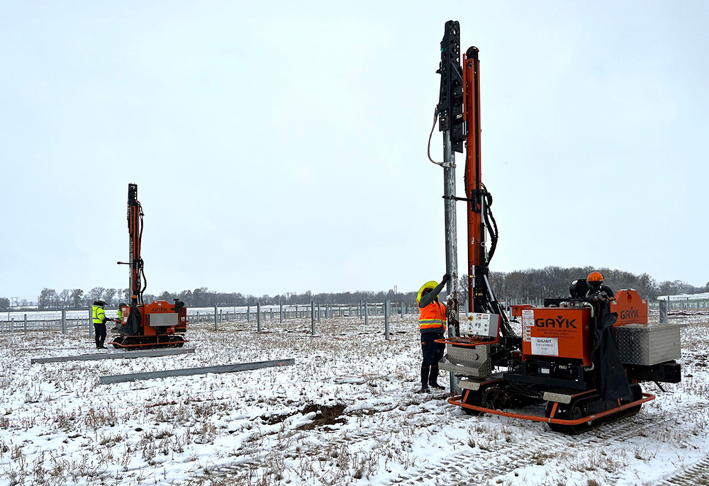 Workers drive piles at a snowy solar tracker project site.